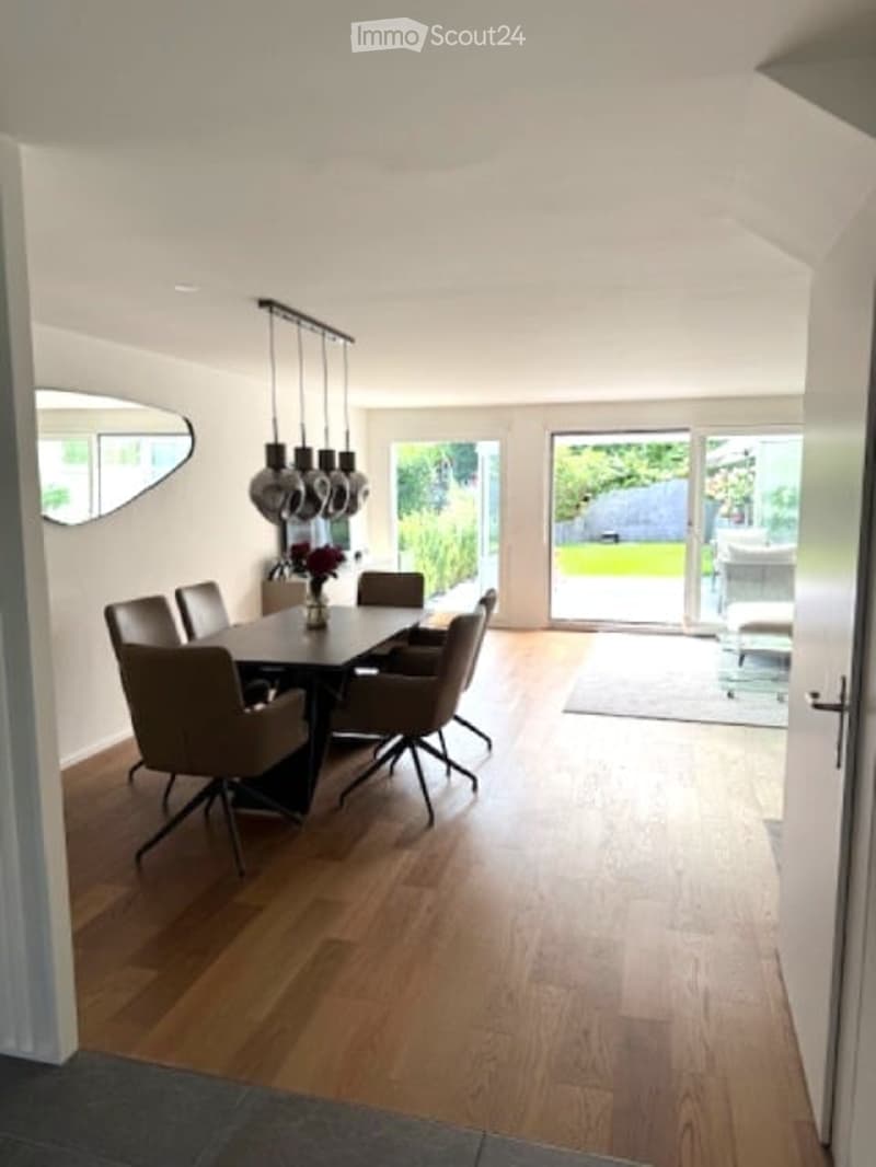 Bright dining room with wooden flooring, a black dining table surrounded by six chairs, pendant lights above, and a large mirror on the wall. A sliding glass door leads to a garden view.