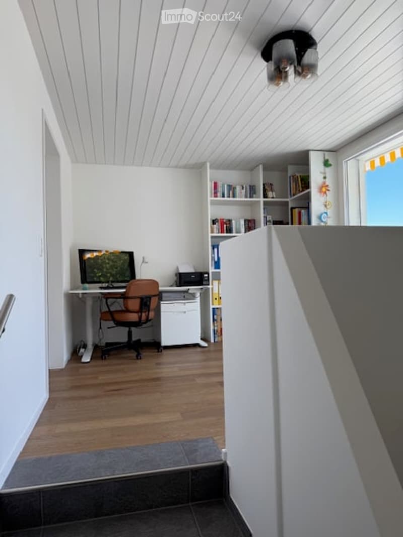 A room with a desk, chair, bookcase, monitor, printer, and other office equipment, wooden floor, white ceiling, wall, and stairs