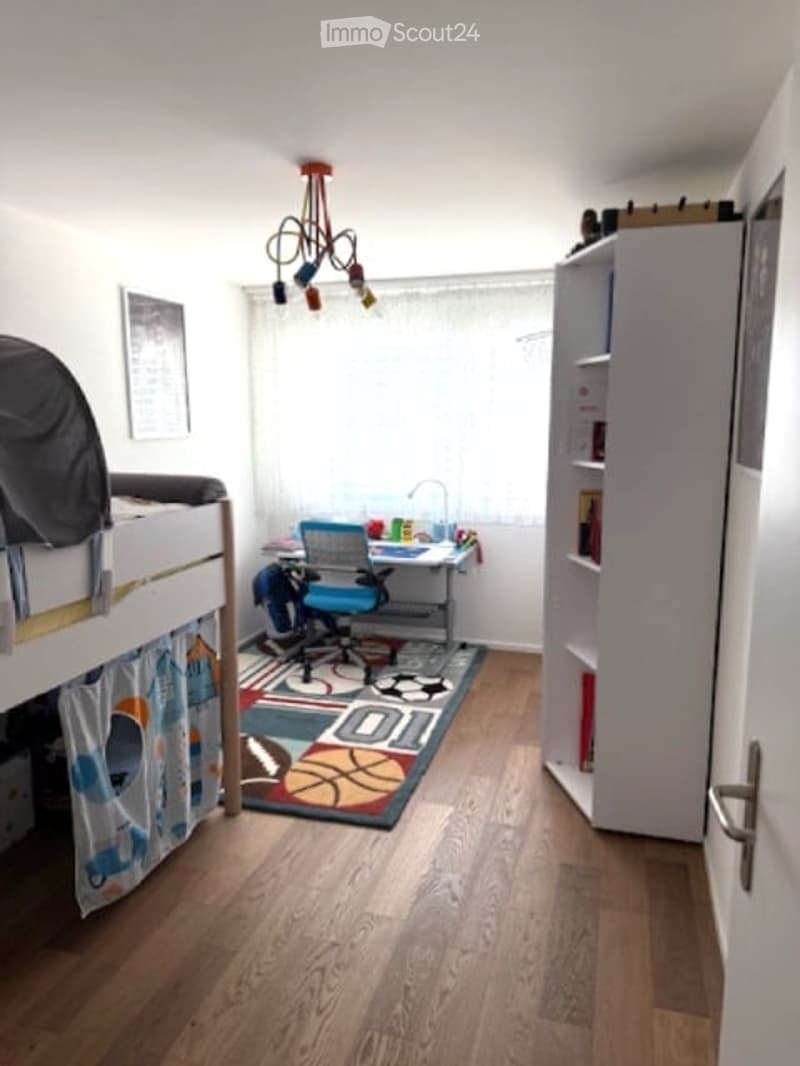 A small, well-lit room with a bunk bed, desk, chair, bookshelf, and a colorful rug on the floor.