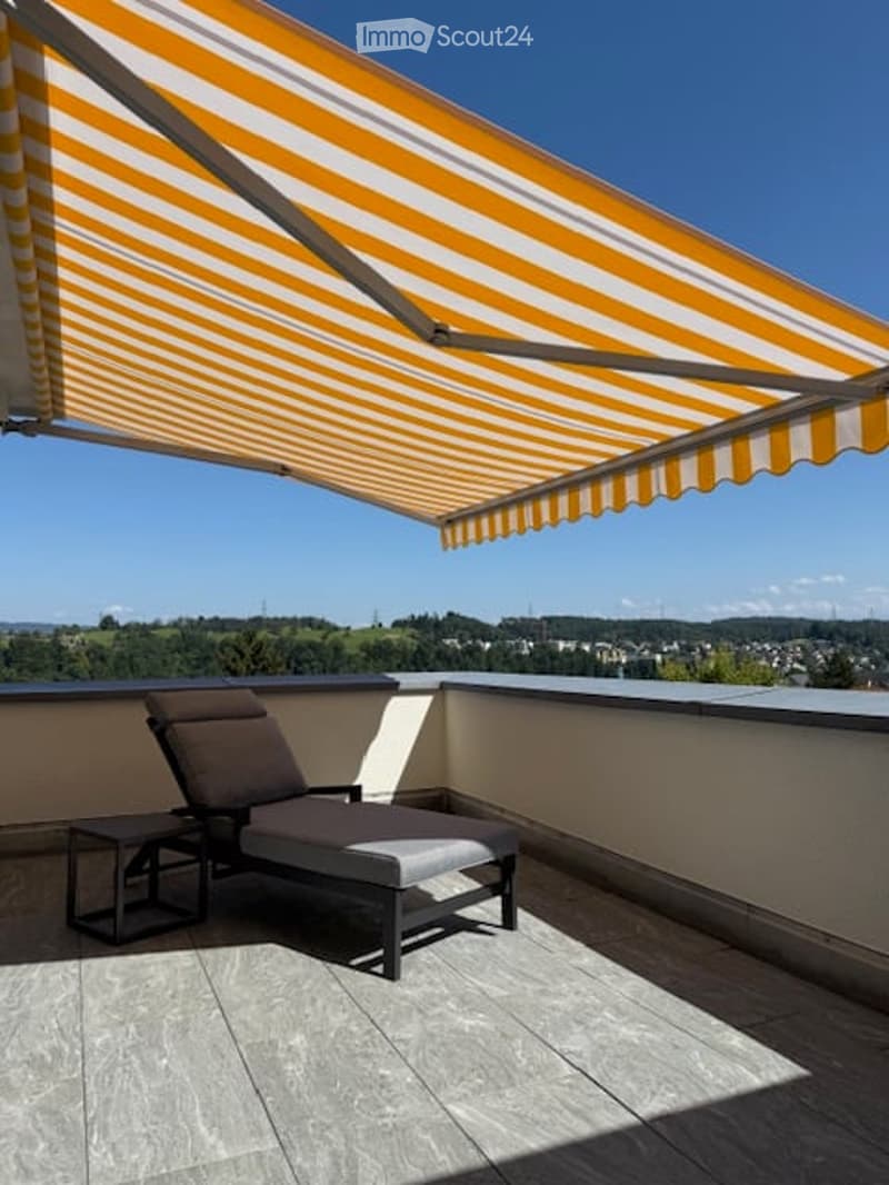 Balcony with awning, lounge chair, table, tiled floor, distant view of trees and houses