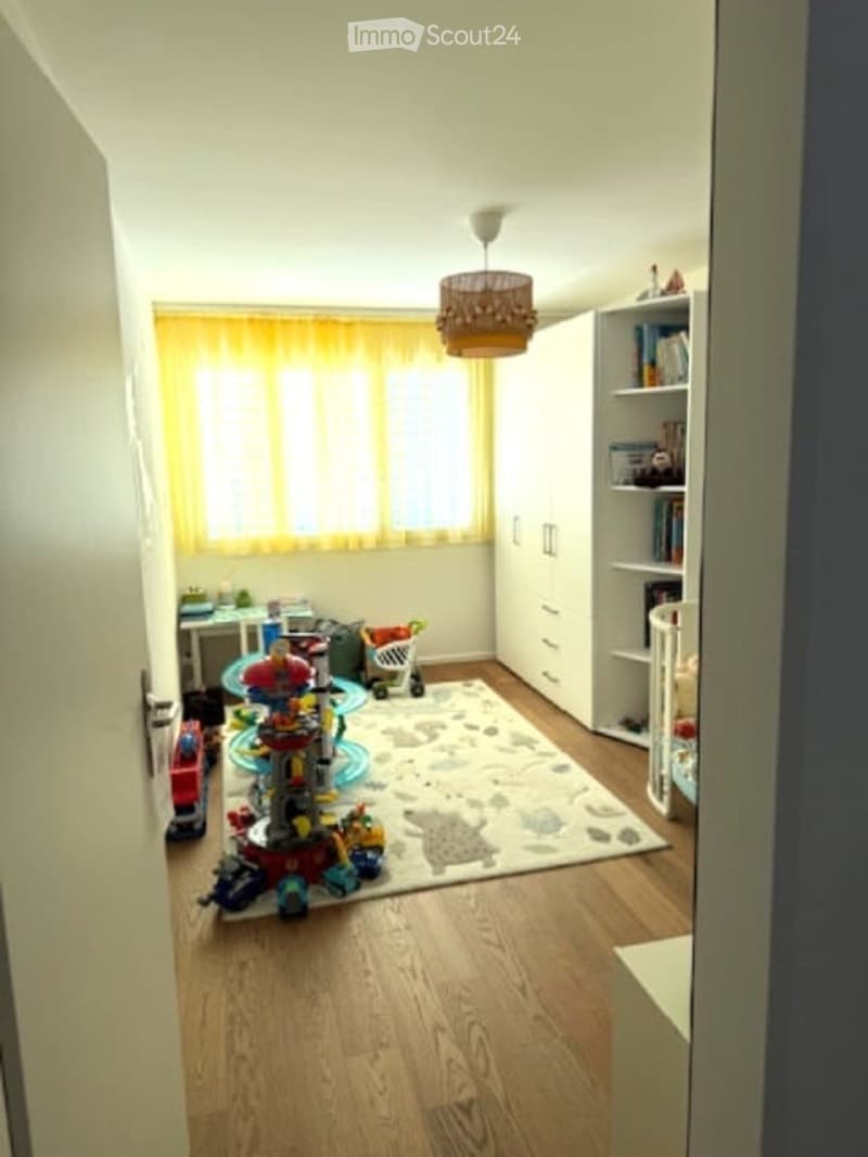 A well-lit children's room with wooden flooring, a large window with yellow curtains, a white wardrobe, a bookshelf, a small table with a chair, a colorful toy structure, a toy car, a toy robot, a toy stroller, and a rug with animal prints.