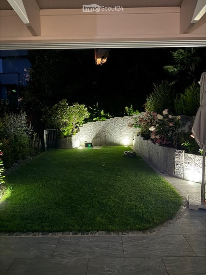 landscaped garden with green lawn, illuminated by spotlights, surrounded by low stone walls and diverse plants