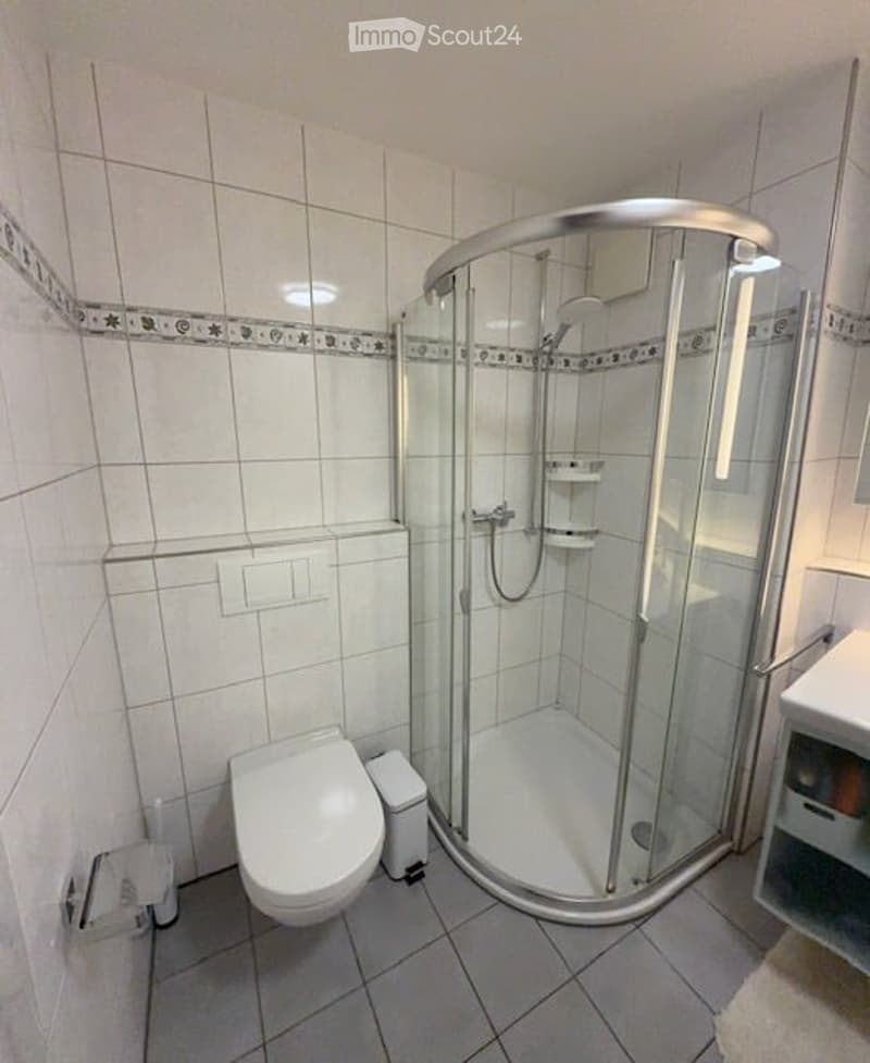 Bathroom with white tiles, glass shower enclosure, toilet, and a small trash bin.