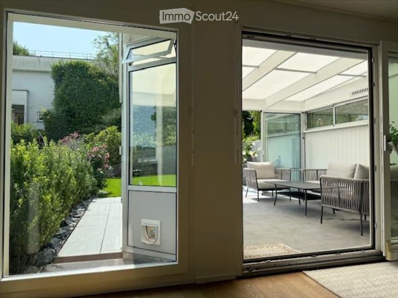 Open sliding glass doors lead to a covered patio area with outdoor furniture, including a sofa, chairs, and a coffee table. The patio is surrounded by a well-maintained garden with hedges and a pathway.