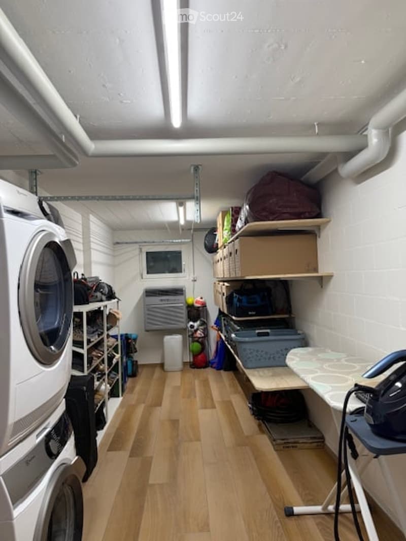 Laundry room with wooden floor, white walls, washing machine, shelves, ironing board, storage boxes, and various items