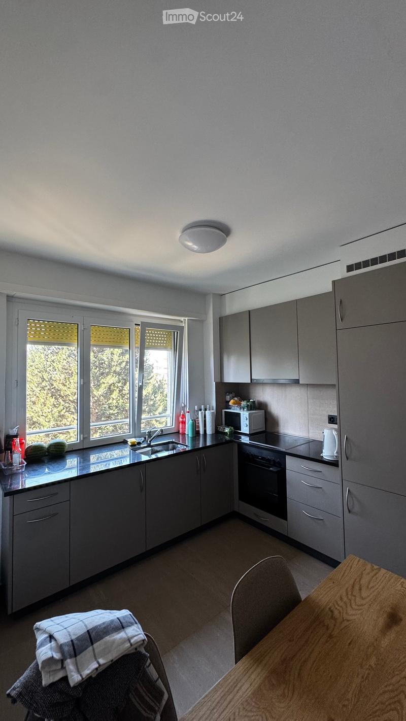 Modern kitchen with grey cabinets, black countertops, and stainless steel appliances. Features a sink, microwave, oven, and a small dining area with a table and chair.