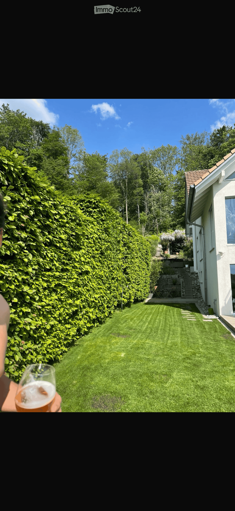 Well-maintained lawn bordered by a tall hedge, with a small patio area and outdoor furniture near the house.