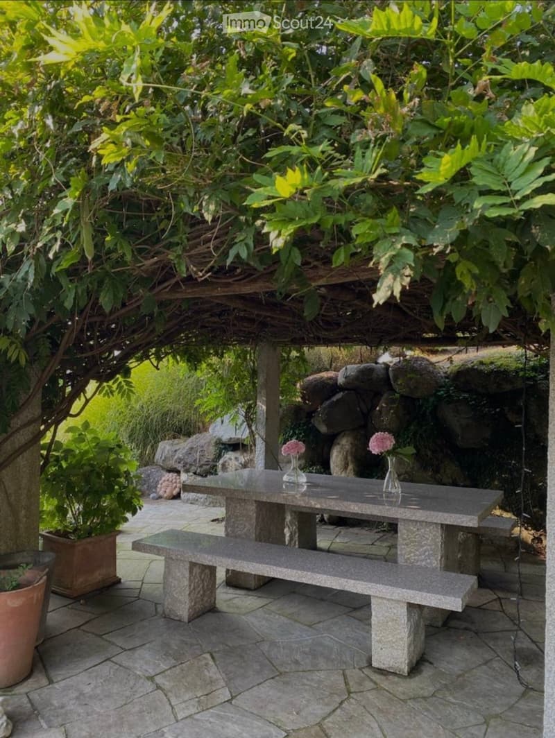 A serene outdoor seating area with a stone table, bench, and two vases with pink flowers, surrounded by lush greenery and large rocks.