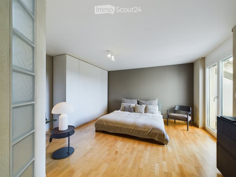Modern bedroom with wooden flooring, large bed with pillows and blankets, nightstand with lamp, chair, and built-in wardrobe.
