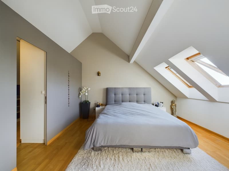 Modern bedroom with a grey upholstered headboard, wooden floor, and skylight windows. Features a nightstand with a lamp, a decorative vase, and a small statue.
