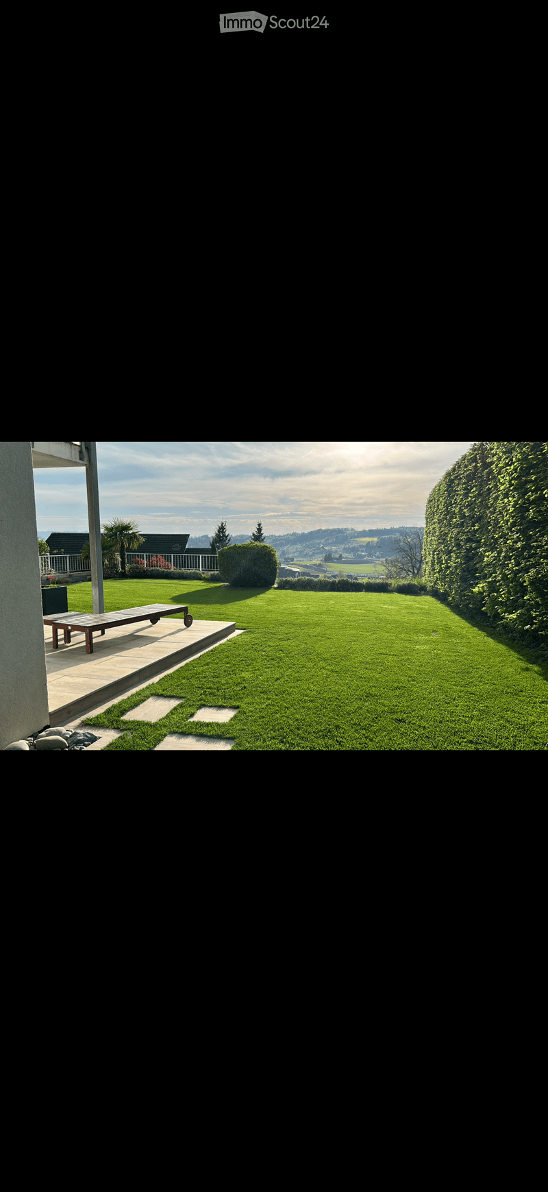 Well-maintained lawn with a wooden bench on a patio, surrounded by hedges and trees, with a scenic view of a valley and mountains in the background.