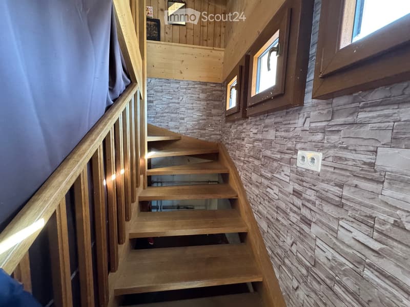 Wooden staircase with a stone-textured wall, small windows, and a wooden handrail.