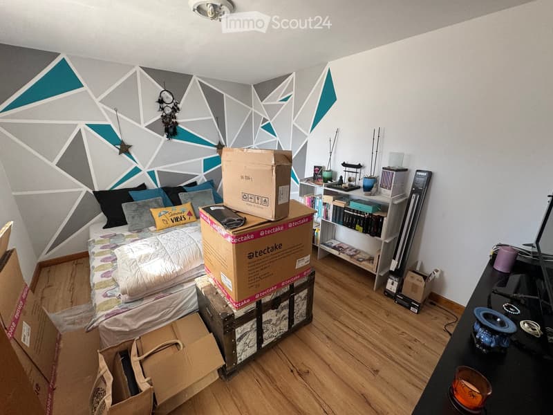 A small bedroom with a bed, pillows, and a colorful blanket. The walls feature a geometric design with grey and teal accents. A shelf holds books and decorative items, and several cardboard boxes are scattered around the room.