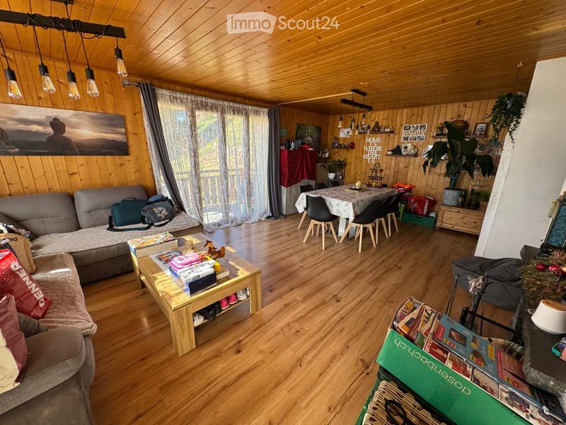 A cozy living room with wooden walls and ceiling, featuring a grey sofa, coffee table, dining area with chairs, and various decorative items including a plant and picture frames.