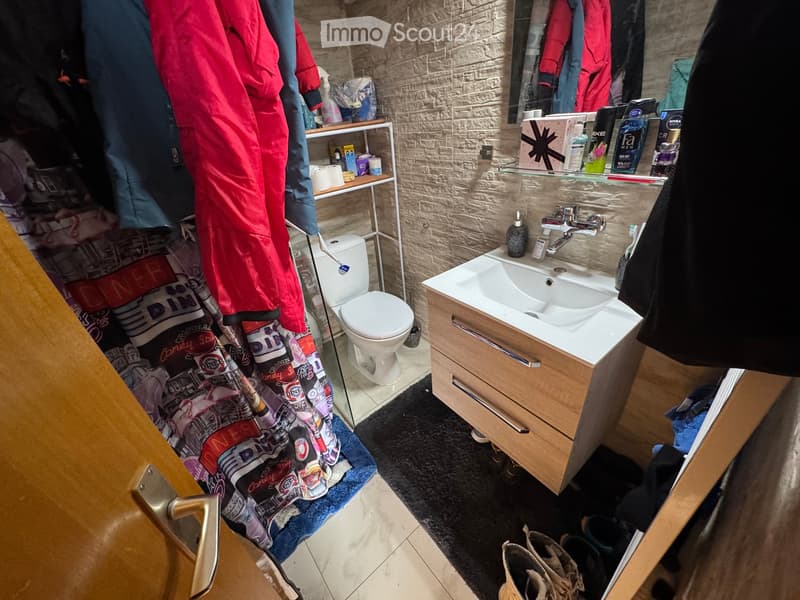 Modern bathroom with a sink, toilet, mirror, and shelf holding personal care items. Clothes and shoes are visible near the door.