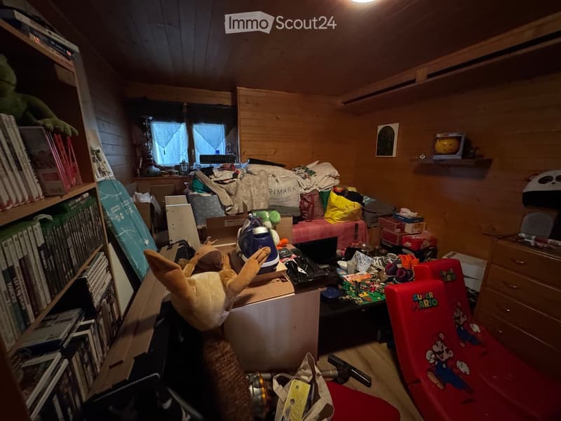 A cluttered room with wooden walls and ceiling, featuring a shelf with books and toys, a table with a laptop, a red chair with a Super Mario design, and various scattered items including a surfboard and a large plush toy.