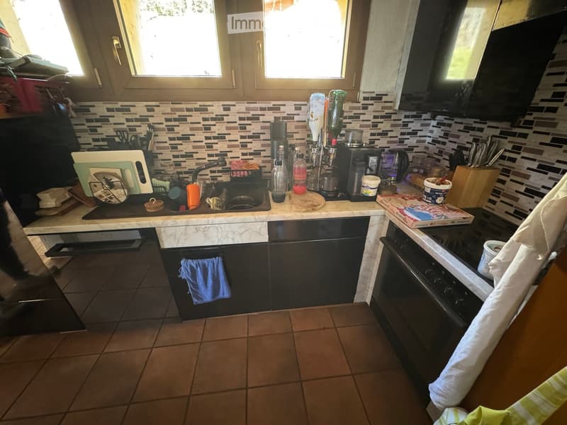 The kitchen features a marble countertop, a sink with a faucet, a coffee maker, a cutting board, a knife block, and various kitchen utensils. The cabinets are dark-colored, and the floor is tiled.