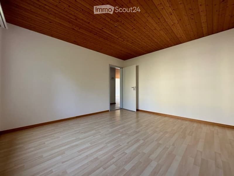 Empty room with wooden flooring and white walls, featuring a wooden ceiling and an open doorway leading to another space.