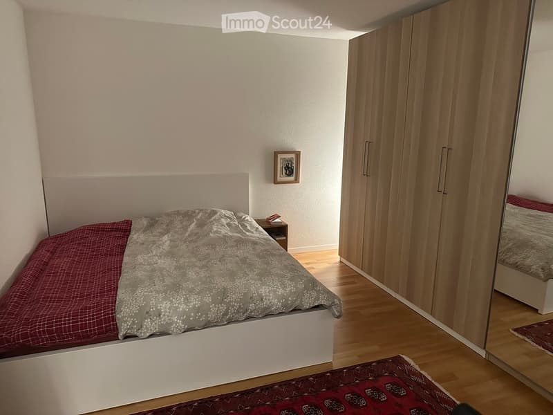 Bedroom with a white bed, red and grey bedding, a wooden nightstand, a large wardrobe, and a mirror. The room has light wooden flooring and a patterned rug.