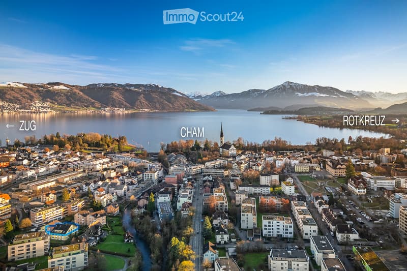 Aerial view of a town with residential and commercial buildings, surrounded by a lake and mountains in the background.