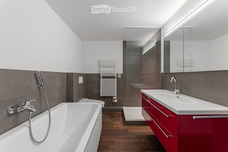 Modern bathroom with a white bathtub, double sink vanity with red cabinets, a large mirror, and a radiator. The space features grey tiled walls and a wooden floor.