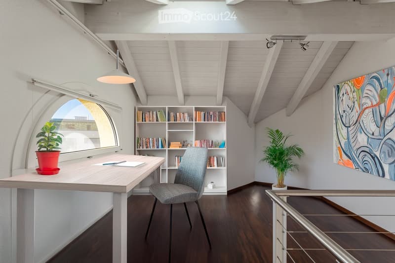 Bright, modern study or home office with wooden flooring, white walls, and exposed wooden beams. Features a large arched window, a wooden desk with a chair, a bookshelf filled with books, and a colorful abstract painting on the wall.