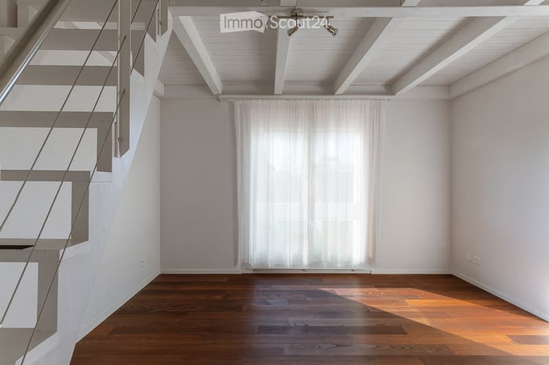 Empty room with wooden flooring, white walls, and a staircase leading upwards. Natural light filters through a large window with white curtains.