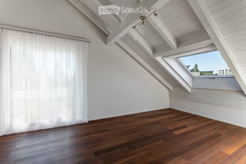Empty attic room with wooden flooring, white walls, exposed wooden beams, skylight windows, and white curtains on one wall.