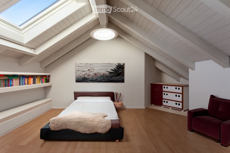 Modern attic bedroom with wooden floors, slanted ceilings, skylight windows, a bed with a fur throw, a red sofa, built-in bookshelf, and a picture frame on the wall.