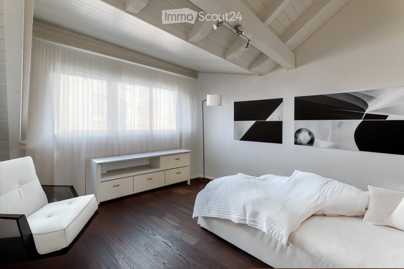 Modern bedroom with a large bed, white bedding, a white leather chair, a sleek white cabinet, and contemporary artwork on the wall. The room features wooden flooring and large windows with sheer curtains.