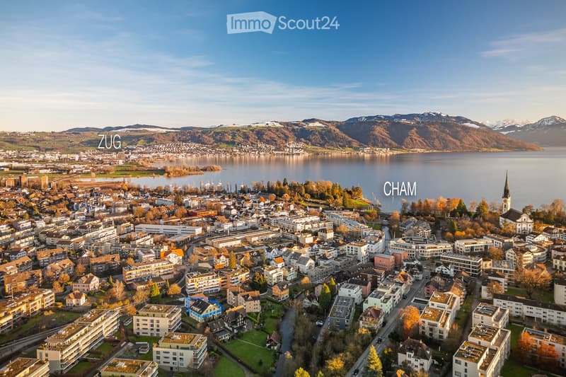 Aerial view of the town of Cham, Switzerland, situated near Lake Zug with surrounding mountains in the background. The town features a mix of residential and commercial buildings, a church spire, and a waterfront area.