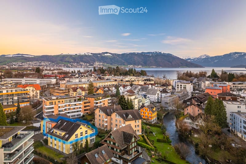 Aerial view of a town with a mix of residential and commercial buildings, surrounded by green spaces, a river, and distant mountains and a lake.