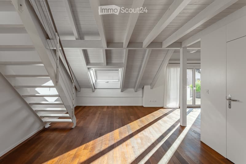 Bright, airy hallway with wooden flooring, white walls, and exposed wooden beams on the ceiling. Features a staircase leading upwards and a large window allowing natural light to flood the space.