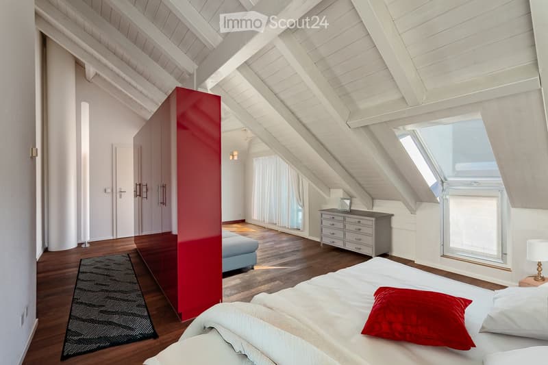 Bright attic bedroom with wooden slanted ceilings, hardwood floors, large windows, and skylight. Features a bed with white linens and red pillows, a nightstand with a lamp, a small sofa, a grey dresser, and a red accent wall.