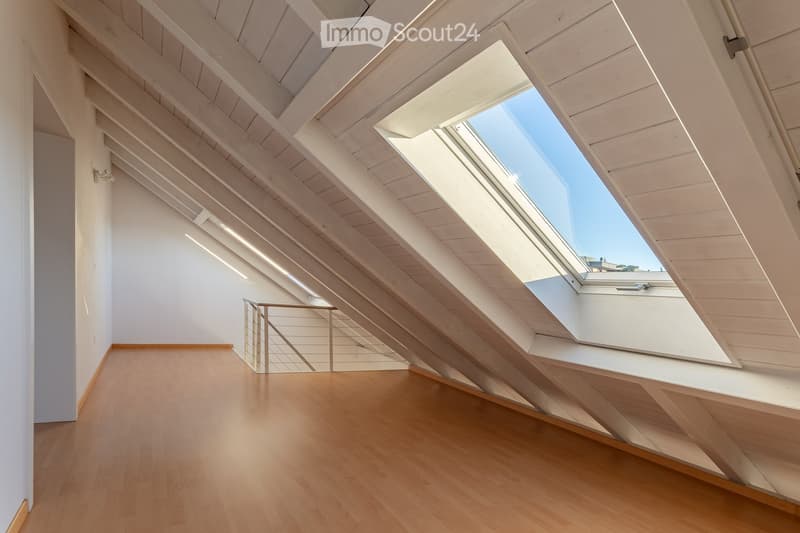 Bright, empty attic space with wooden floors, white walls, exposed wooden beams, and skylight windows allowing natural light.