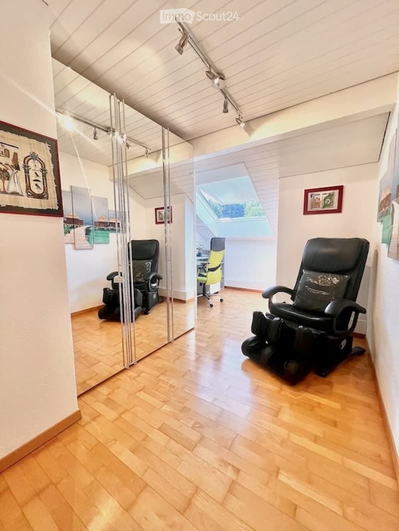 A modern, well-lit room with wooden flooring, large mirror, and two massage chairs. Features a skylight providing natural light and a small desk area.