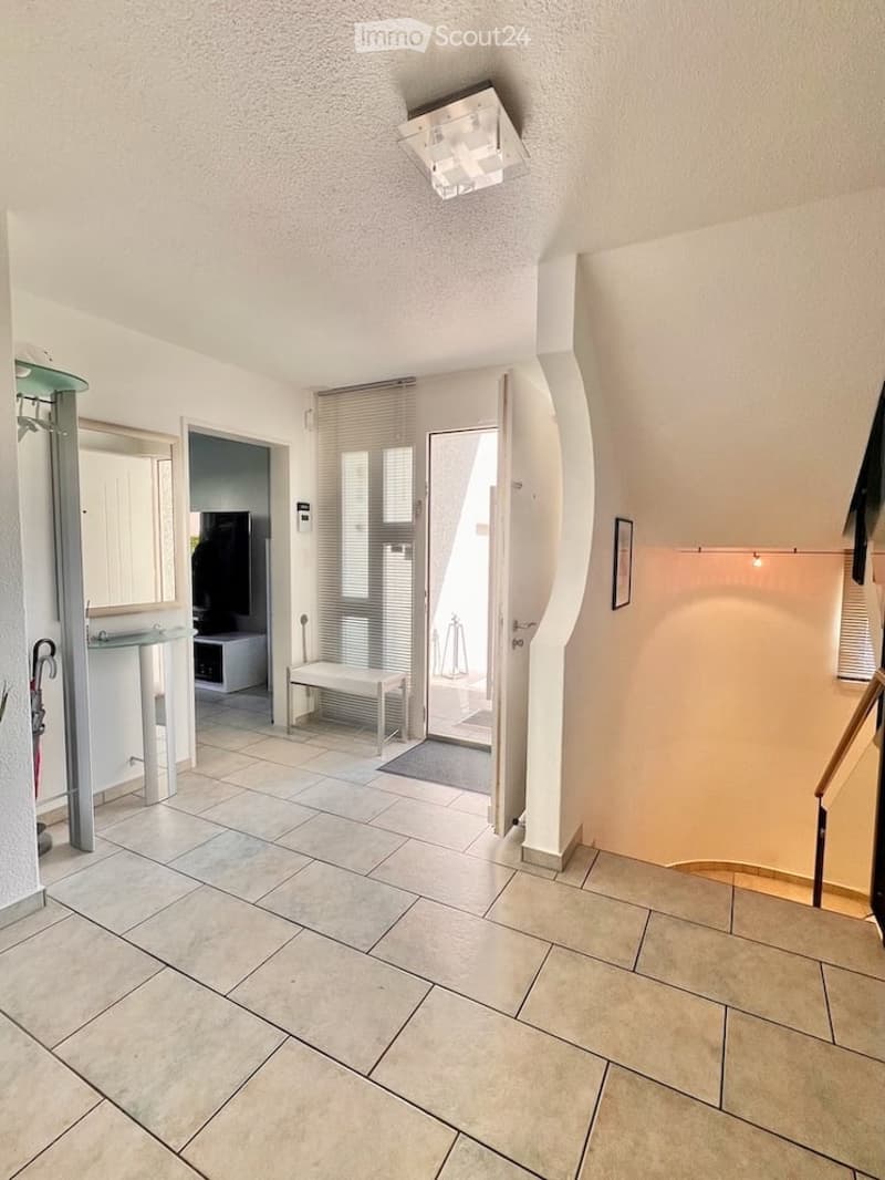 Bright hallway with tiled flooring, white walls, and a modern ceiling light fixture. Features an open doorway leading to another room, a small bench, and a glass console table with a mirror.