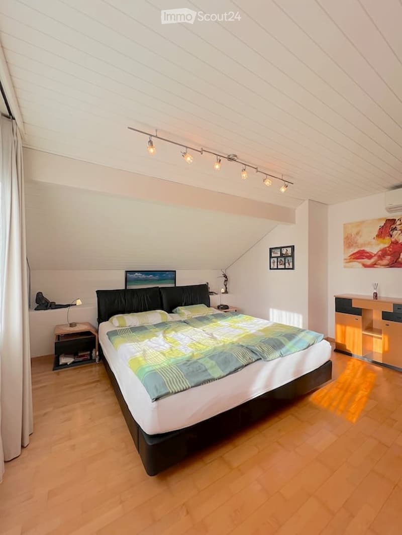 Bright bedroom with wooden flooring, large bed with colorful bedding, nightstand with lamp, desk with chair, and artwork on walls.