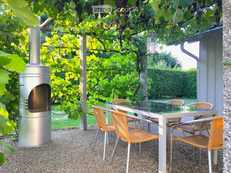 Outdoor patio area with a glass-top dining table, six wicker chairs, and a modern metal wood-burning stove. The space is surrounded by lush greenery and trees, creating a serene and private atmosphere.
