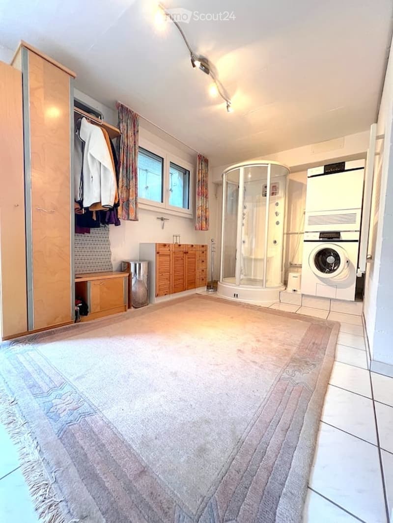 Well-lit room with a large rug, a wardrobe with hanging clothes, a shower stall, a washing machine, and wooden cabinets.