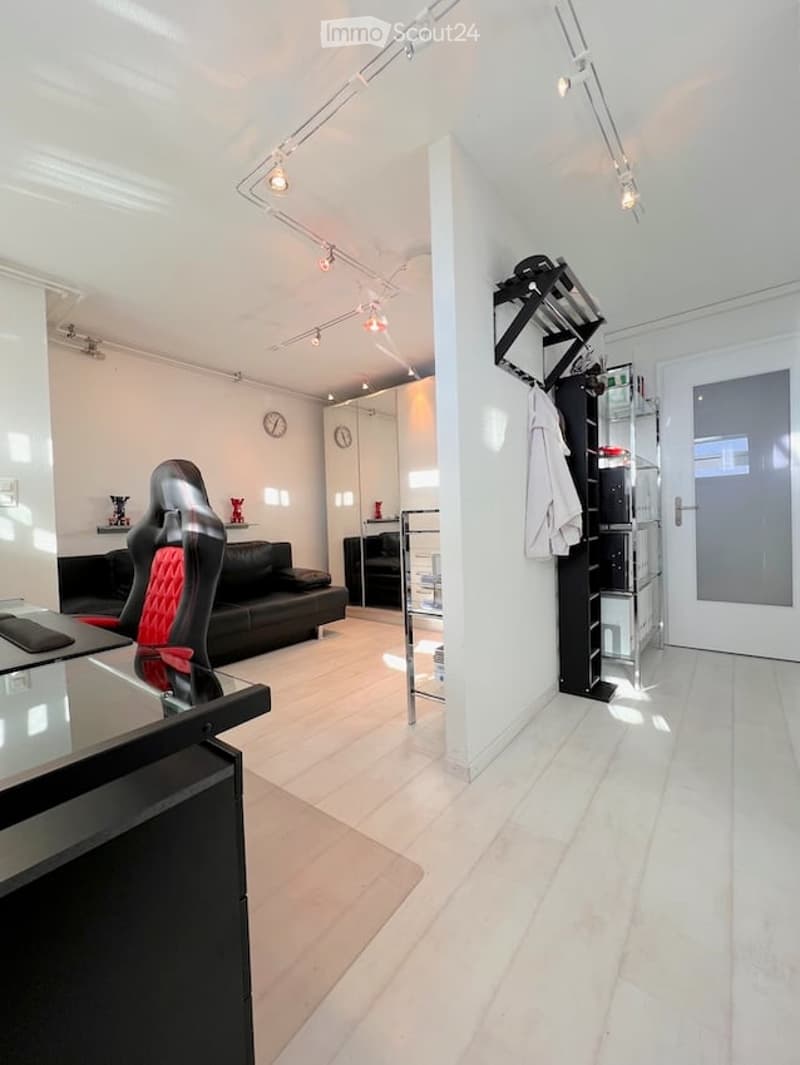 Modern, minimalist living room with white walls, light wooden flooring, and track lighting. Features a black leather sofa, red accent chair, glass coffee table, built-in cabinetry, and a small shelf with decorative items.