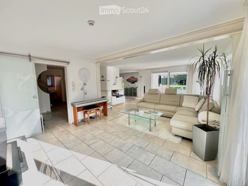 Bright living room with a beige sectional sofa, glass coffee table, piano, and a potted plant. The space features large windows and a modern design.