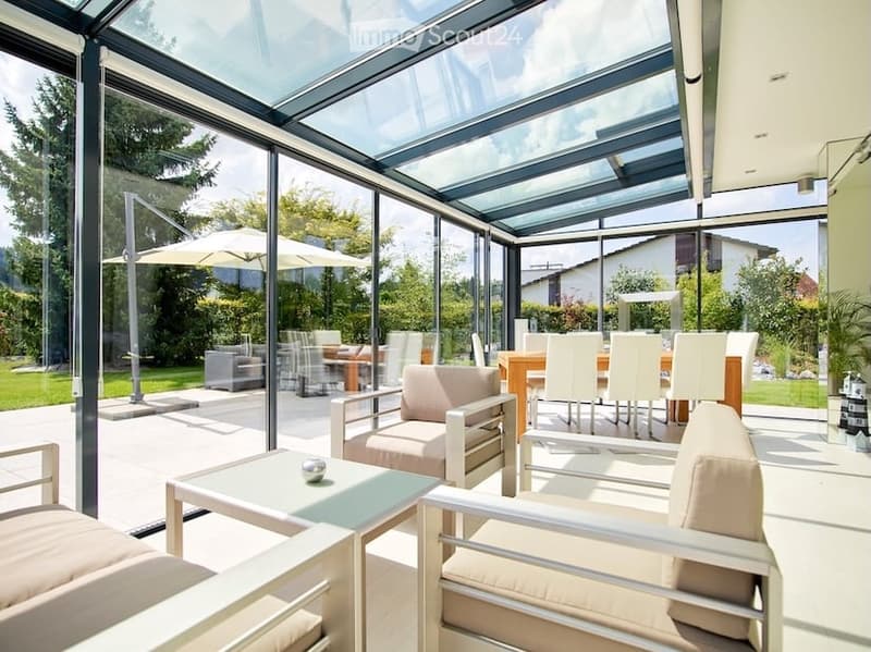 Bright, modern indoor-outdoor living space with large glass walls and a retractable roof, featuring a sofa, chairs, a coffee table, and outdoor dining area with umbrellas.