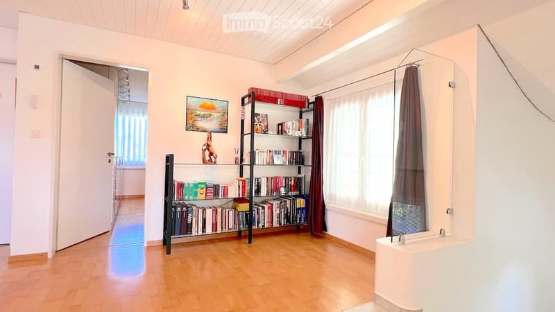 A well-lit room with wooden flooring, a large window with curtains, a bookshelf filled with books, a picture frame on the wall, and a small bathtub in the corner.