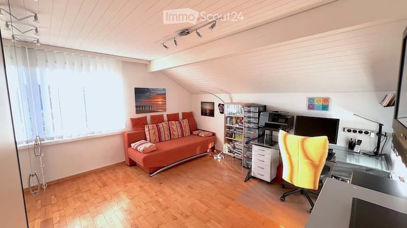 Bright room with wooden flooring, large window with white blinds, orange sofa with colorful cushions, desk with chair, bookshelf, printer, and screen.