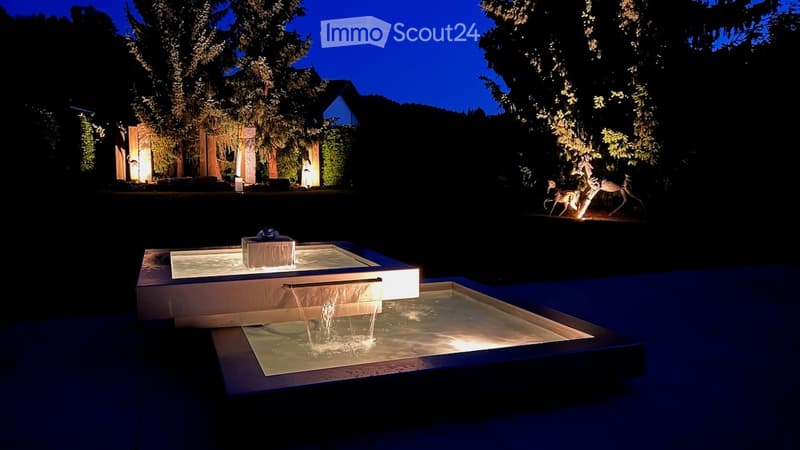 Nighttime view of a modern outdoor water feature with illuminated steps and a small fountain, surrounded by trees and a house in the background.