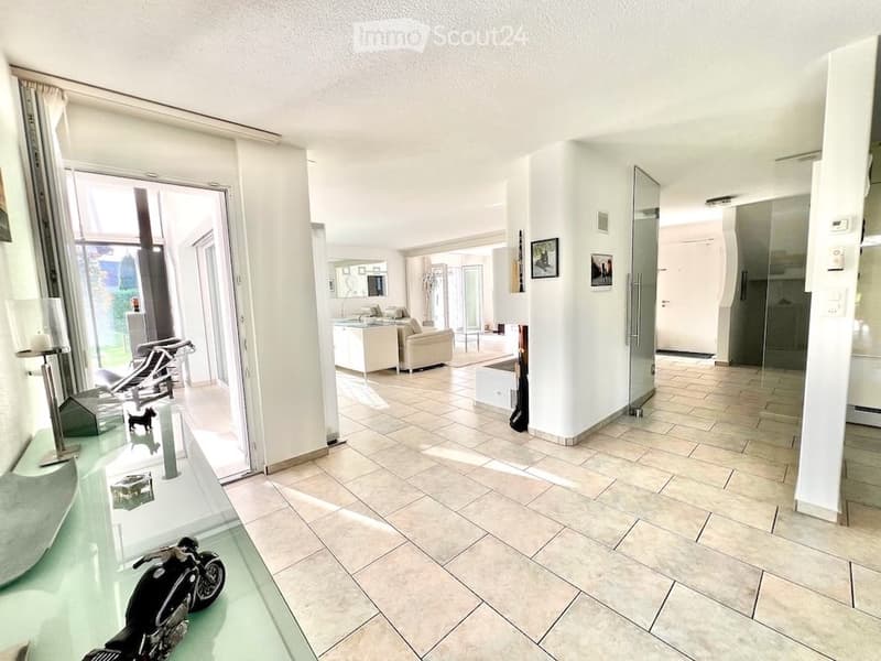 Bright, modern hallway leading to a living area with a sofa, coffee table, and chairs. The space features large windows, tiled flooring, and decorative elements like lamps and artwork.