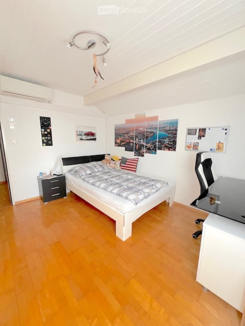 Modern bedroom with wooden flooring, white walls, a single bed with a plaid blanket, a nightstand, a desk with a chair, and several picture frames on the walls.
