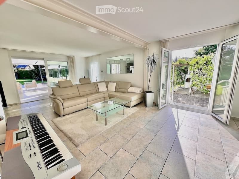 Modern living room with large sliding glass doors leading to a patio, beige sectional sofa, glass coffee table, piano, and a potted plant near the entrance.