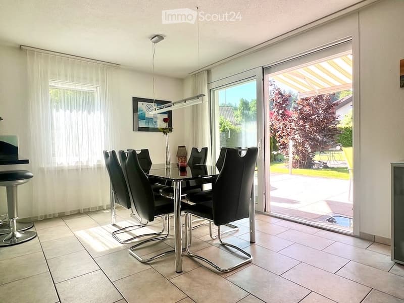 Modern dining room with a glass dining table, black chairs, a stool, and a lamp. Large windows and sliding glass doors lead to an outdoor area.